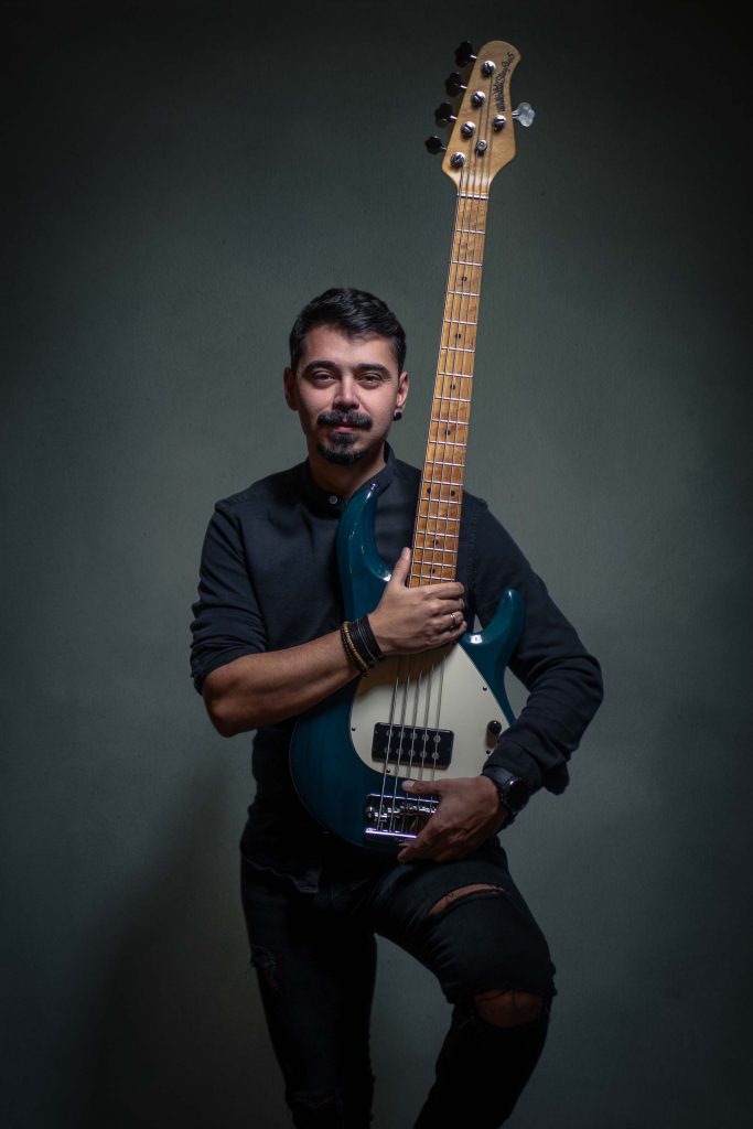 Mladi muškarac, basista, u crnoj majici i farmerkama sa rupicama, drži bas gitaru.  Fotografija je snimljena u studiju u Novom Sadu, za promociju muzičkog benda.  Profesionalno osvetljenje i kompozicija naglašavaju instrument i izraz lica basiste.