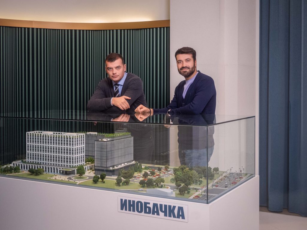 Profesionalno korporativno fotografisanje menadžmenta kompanije Inobacka u Novom Sadu.  Na slici su dva člana menadžmenta u kancelarijama kompanije.  Fotografija je snimljena u modernom ambijentu, sa fokusom na predstavljanje kompanije.  U prvom planu se nalazi model budućeg poslovnog kompleksa Inobacka.  Kvalitetno osvetljenje i kompozicija fotografije ističu važnost i profesionalnost kompanije.