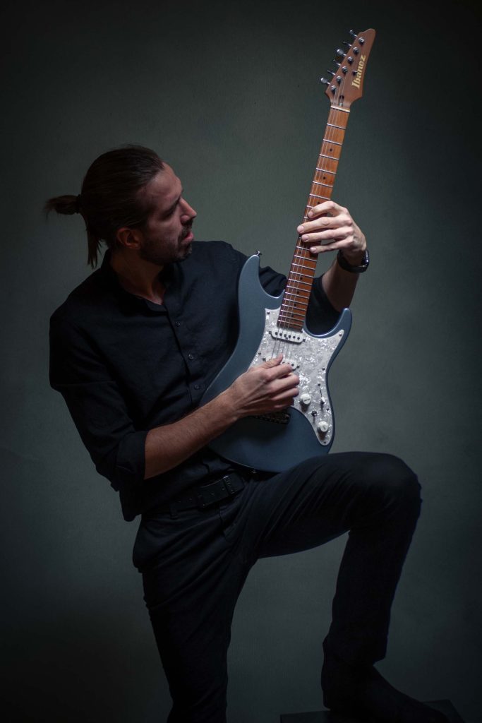 Muškarac u crnoj košulji i pantalonama, svezan konjski rep, svira električnu gitaru Ibanez.  Profesionalna fotografija u studiju u Novom Sadu, namenjena promociji muzičkog benda.