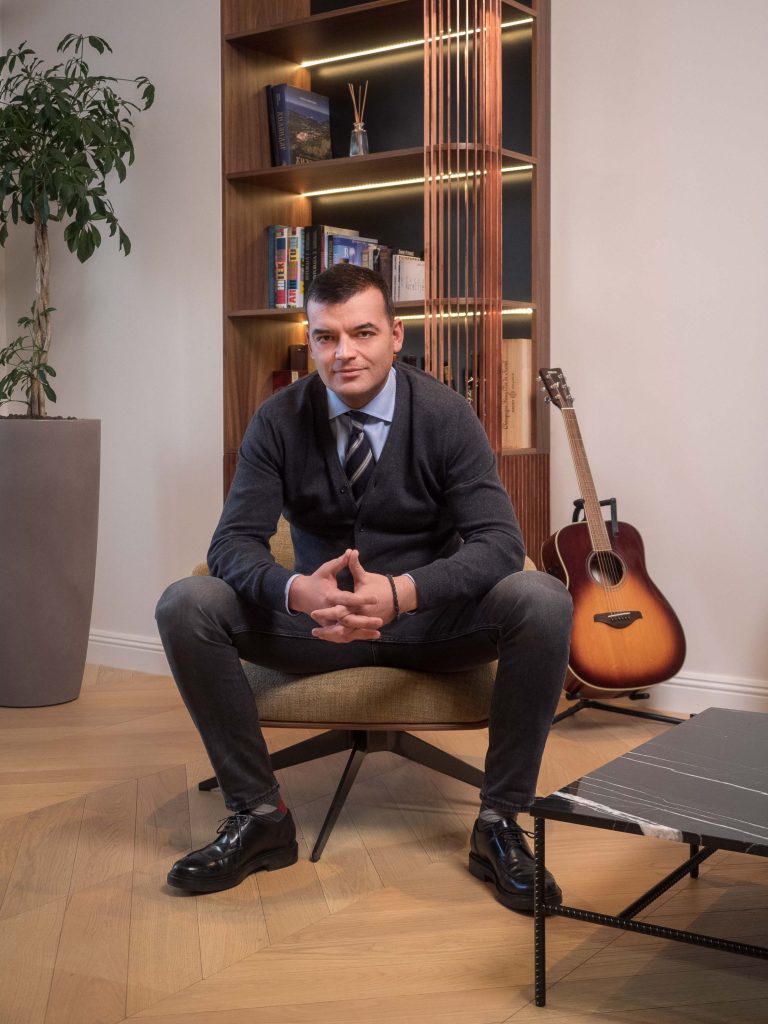 Profesionalno fotografisanje menadžmenta kompanije Inobacka u Novom Sadu.  Na slici je muškarac u poslovnom odelu, sedeći u kancelariji.  Moderan enterijer sa drvenim elementima, elegantnim nameštajem i detaljima kao što su biljke i gitara.  Fotografija je snimljena u kancelarijama kompanije, što naglašava profesionalni i ugledan ambijent.  Idealno za korporativnu prezentaciju i promociju Inobacke.