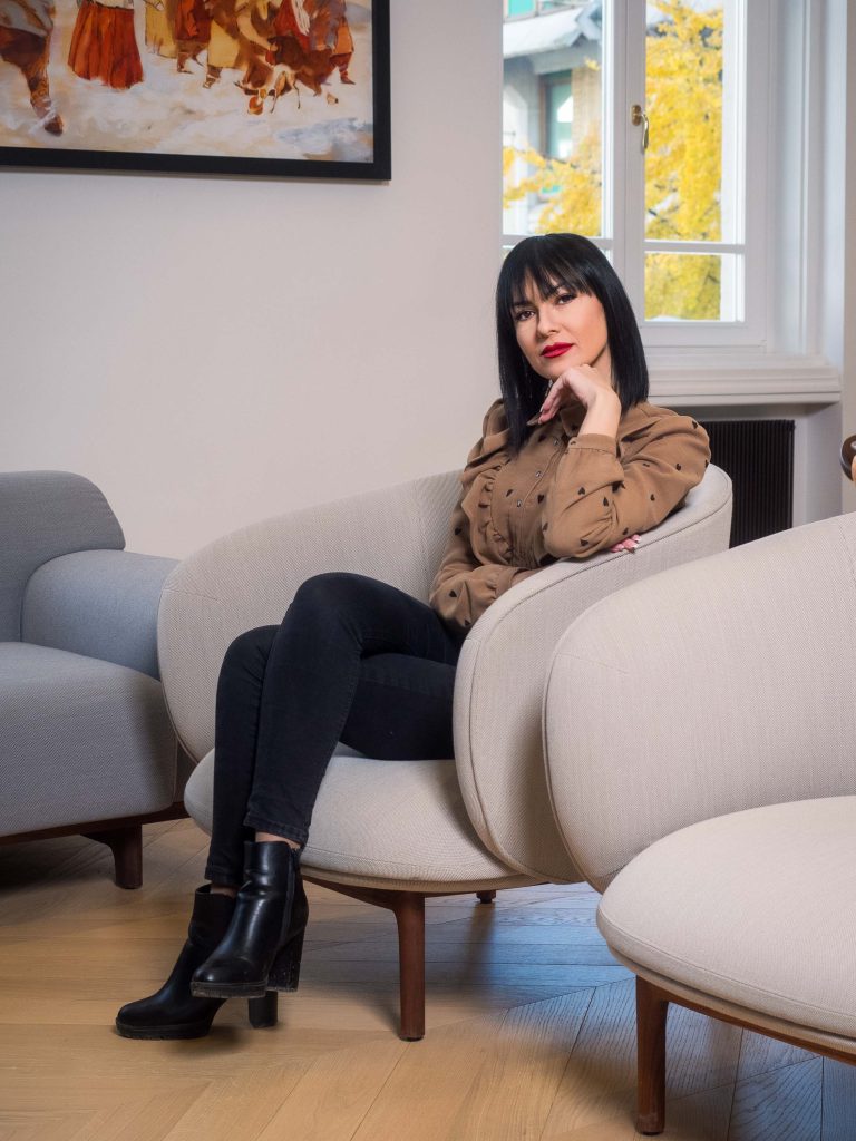 Na fotografiji je žena, članica menadžmenta kompanije Inobacka, u kancelarijama u Novom Sadu.  Sedi u elegantnom, bež kauču sa drvenim nogama.  Nosi crne čizme na visokom potpetici i smeđu košulju sa crnim šarama u obliku srca.  Ambijent je moderan i udoban, sa bež tapacirungom i drvenim podom u obliku šahovnice.  Na zidu je velika slika.  Fotografija je profesionalno osvetljena i prikazuje dobar kvalitet.