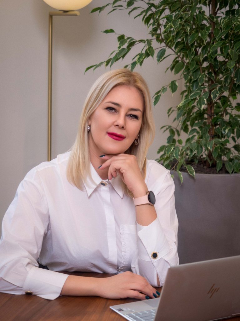 Profesionalno fotografisanje menadžmenta kompanije Inobacka u Novom Sadu.  Na slici je žena u beloj košulji, elegantnog izgleda, sa osmehom.  U pozadini su kancelarijski prostor i biljka.  Fotografija je snimljena u kancelarijama kompanije, što naglašava profesionalni i uredan ambijent.