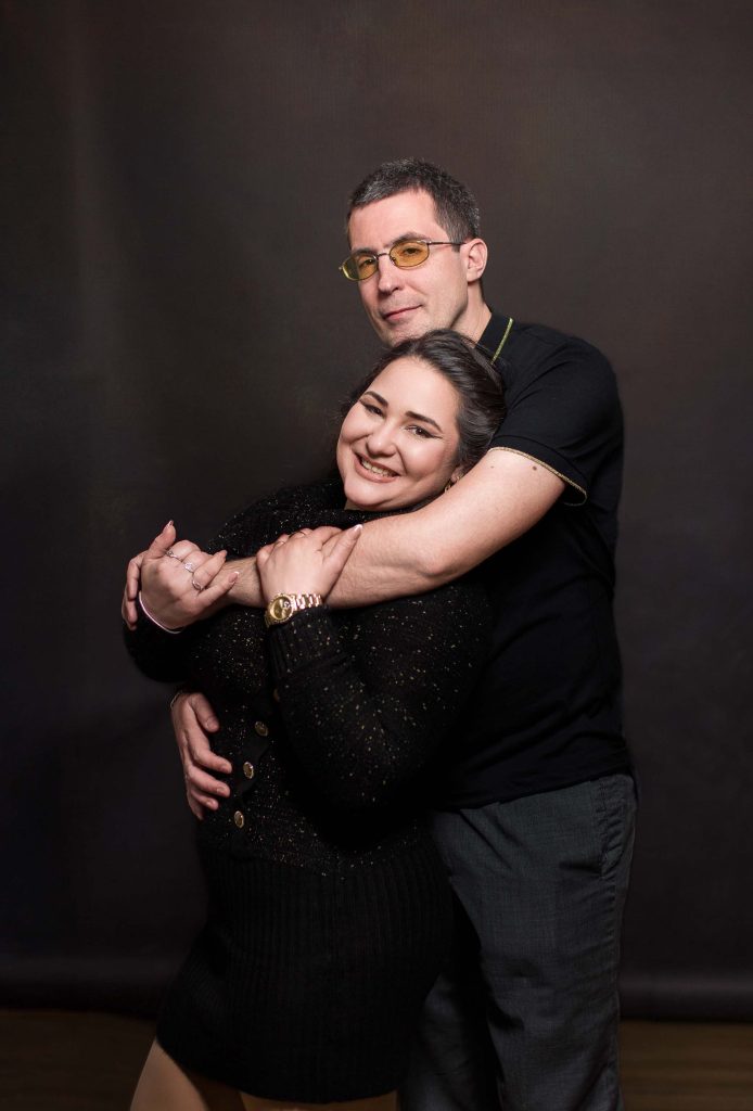 Zaljubljeni par u zagrljaju, ovekovečen u mom studiju u Novom Sadu. Ona, u elegantnoj crnoj haljini, blista od sreće, dok on, sa osmehom na licu i naočarima, nežno grli svoju dragu. Njihova povezanost i ljubav su očigledne, a toplina koju emituju ispunjava čitavu fotografiju. Uvek mi je zadovoljstvo da beležim ovakve trenutke. Milan Stojanović, profesionalni fotograf.